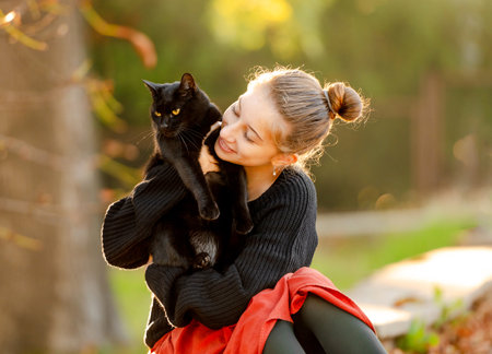 Pretty girl in red skirt hugging black cat outdoors at street with autumn leaves. Beautiful model teenager sitting with feline animal at parkの写真素材