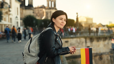 Young Woman Holds German Flag In Hand, With Blurred City Background In Autumnの写真素材