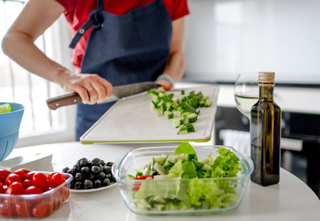 Girl Pours Chopped Cucumbers Into Bowl For Greek Salad At Home In Bright Kitchenの写真素材