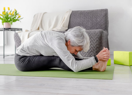 Elderly Athletic Woman Bends While Sitting On Yoga Mat At Home On Floor In Bright Room With Flowersの写真素材
