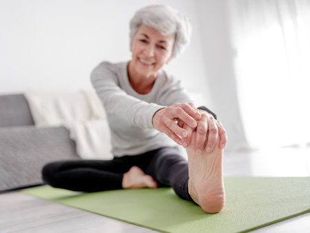 Elderly Athletic Woman Stretches To Her Foot While Sitting On Yoga Mat, Exercising At Home On Floor In Bright Room With Flowersの写真素材