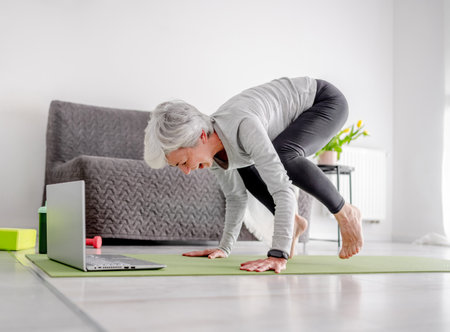 Pleasant 70-Year-Old Woman Performs Complex Exercises With Trainer Through Laptop, Doing Yoga Online At Homeの写真素材