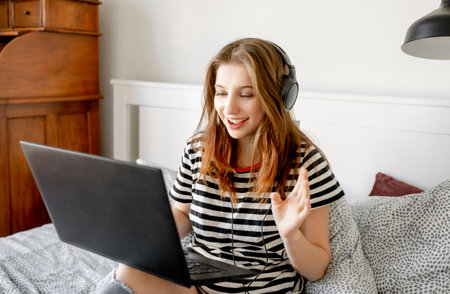 Pretty girl with headphones and laptop in bed during online learning webinar. Beautiful teenager making video call with computer at bedroomの写真素材