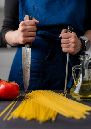 Large Kitchen Knife And Sharpener In Female Hands Are In Close-Up Over Table With Spaghetti, Tomatoes, Spices, And Basilの写真素材