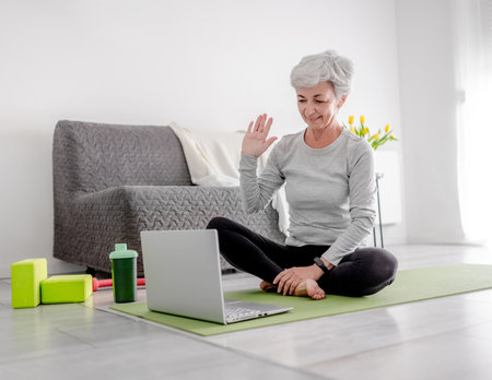 Pleasant 65-Year-Old Woman Greets Her Trainer Through Laptop For An Online Sport Session At Home On A Yoga Matの写真素材