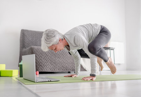 Pleasant 70-Year-Old Woman Performs Complex Exercises With Trainer Through Laptop, Doing Yoga Online At Homeの写真素材