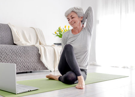 Pleasant 70-Year-Old Woman Exercises With Trainer Through Laptop, Doing Sports At Home On Yoga Matの写真素材