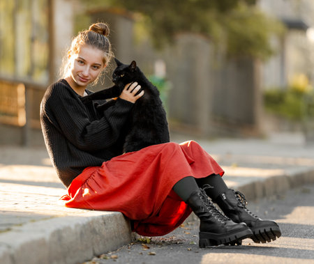 Pretty girl in red skirt hugging black cat outdoors at street with autumn leaves. Beautiful model teenager sitting with feline animal at parkの写真素材