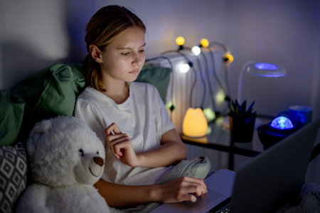 Cute Little Girl In Bed Browses The Internet On Laptop Before Sleep With Lovely Teddy Bearの写真素材
