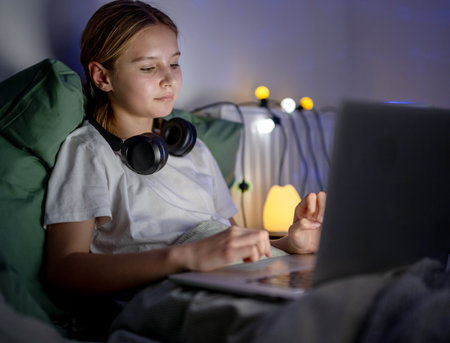 Cute Little Girl Browses The Internet And Social Media and Searching on laptop In Bed Before Sleepの写真素材