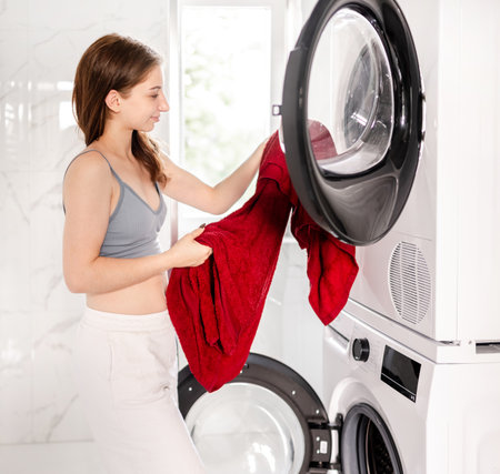 Sweet Young Woman Loading Clothes Into A Dryer In The Bathroomの写真素材