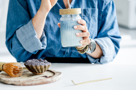 Woman Drinks Blue Iced Matcha Latte In The Kitchen Near Accessories For Its Preparationの写真素材