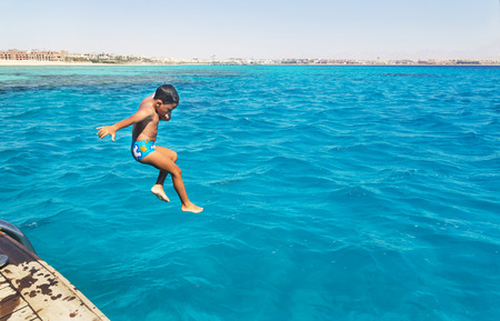 Little arabic boy jumping into the blue water of the sea with yachts.のeditorial素材
