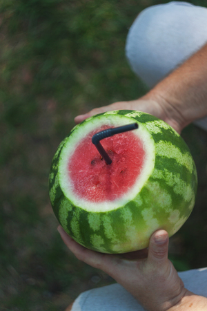 A young man holds a whole watermelon with a straw for drinking, a cocktail. The concept of healthy nutrition and vegetarianism.の写真素材