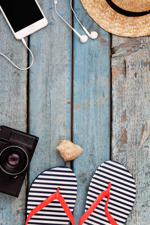 Still life of different items for relaxing on the beach, rubber flip-flops, hat, camera, phone, sunglasses, headset on a wooden blue background. Concept of a youth trip, rest on the beach with perfumed gadgets.の写真素材