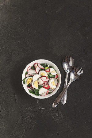Vegetable vegan salad of ramson, radish, green onions and boiled eggs in a white plate on a dark concrete background, in retro treatment. Close-up, top view, flatlay. The concept of healthy nutritionの写真素材