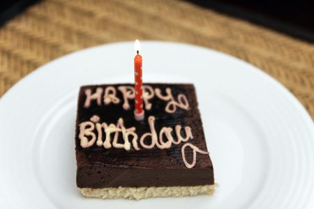 Small square festive chocolate cake with the inscription happy birthday and one candle on a white...
