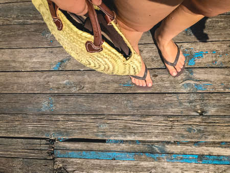 Top view, photo of legs in beach flip-flops and with a straw bag in hand on a wooden old floor. Photos on vacation, beach, summer.の写真素材