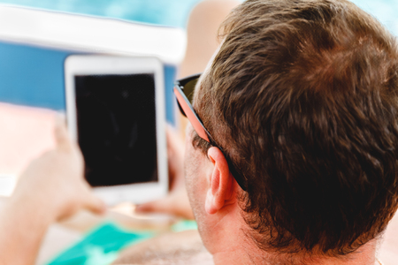 A young man in sunglasses holds a tablet in his hands. Freelancer job concept on the beach and vacation.の写真素材