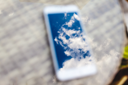 Reflection of blue sky with clouds in the phone-smartphone, which lies on a bright wicker table against the background. Blur and abstractionの写真素材