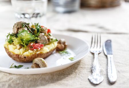 Whole baked potato with mushrooms, tomatoes and vegetables. Serving on a white plate on a light rustic fabric background. Close-up. Top view. Copy spaceの写真素材