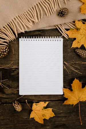 White blank notebook and pen on a dark background with a scarf, plaid and a cup, autumn yellow leaves and pine cones around. View from above. flat lay, in blur. Copy space.の写真素材