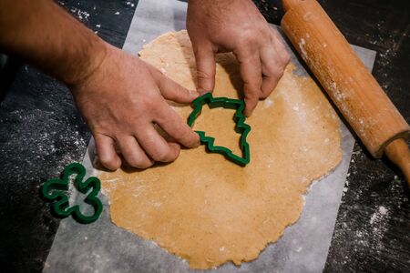 male hands cut out gingerbread cookie in the form of a snowflake, Christmas tree, a man from raw dough on parchment baking paper on a dark background. View from above. save space.の写真素材