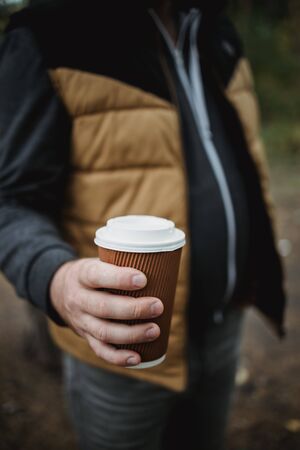 A man holds a takeaway cardboard glass with tea coffee on a background of autumn leaves. Warm autumn atmosphere, soft focus. Copy space.の写真素材