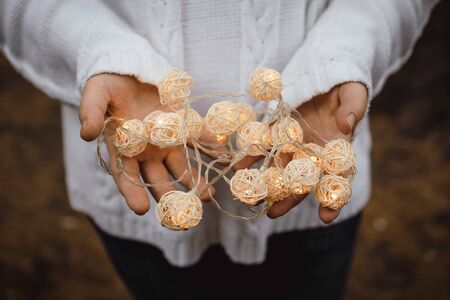 lights in the palms. Female hands hold a garland with wicker round balls. Girl in a white sweater with Christmas lights in her hands, warm New Year mood, soft focus.の写真素材
