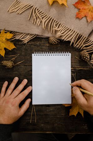 Human hands with a cup of coffee and scarf at wooden table with notebook and pen and autumn leaves. Human hands with a cup of coffee and scarf at wooden tableの写真素材