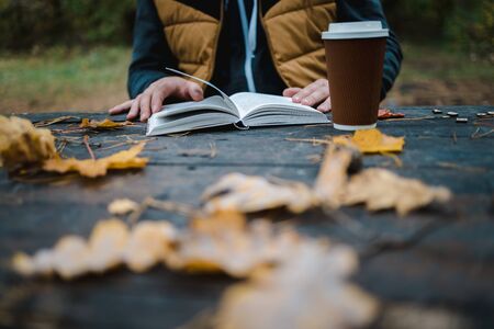 male female reads a book in autumn park drinks takeaway coffee coffee. Close-up over shoulder,...