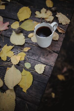 On a old wooden table in the autumn park is a cup with tea and coffee, scattered yellow leaves and pine cones. Top view, in blur. Autumn warm dark mood, soft focusの写真素材