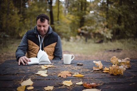a young man sits in an autumn park at an old wooden table, drinks tea and reads a book. Warm dark autumn atmosphere, soft focus. In blur, copy space.の写真素材