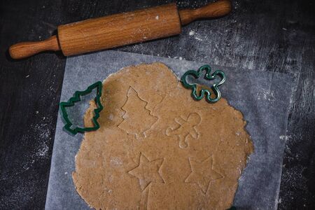 cut out gingerbread cookie in the form of a Christmas tree, star, little man, hearts from raw dough on parchment baking paper on a dark background. Top view. save space.の写真素材