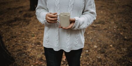 young woman in a white sweater holds a cup of tea with coffee on a background of yellow autumn leaves. Warm autumn atmosphere, soft focus. Copy space.の写真素材