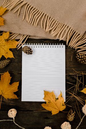 White blank notebook and pen on a dark background with a scarf, plaid and a cup, autumn yellow leaves and pine cones around. View from above. flat lay, in blur. Copy space.の写真素材