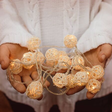 lights in the palms. Female hands hold a garland with wicker round balls. Girl in a white sweater with Christmas lights in her hands, warm New Year mood, soft focus.の写真素材