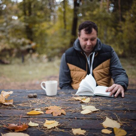 a young man sits in an autumn park at an old wooden table, drinks tea and reads a book. Warm dark autumn atmosphere, soft focus. In blur, copy space.の写真素材