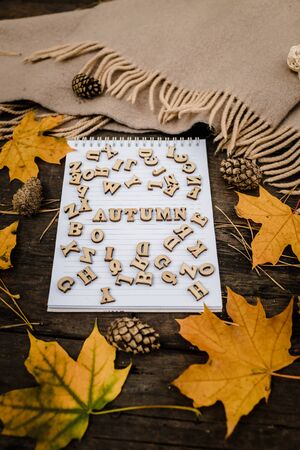 White blank notebook and pen with wooden letters the word autumn on a dark background with a scarf, plaid and a cup, autumn yellow leaves and pine cones around. View from above. flat lay, in blur. Copy spaceの写真素材