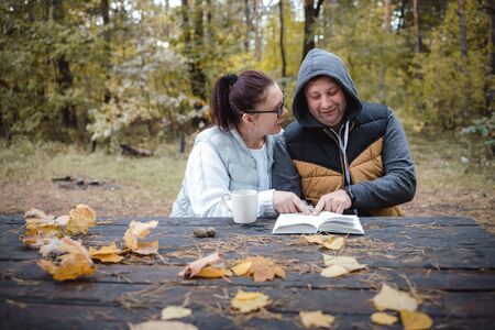 Handsome loving couple sitting on the bench in the autumn park reading a book, having fun together. Love and tenderness. Relationships, friendships, education, lifestyle concept.の写真素材
