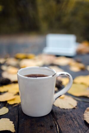 On a old wooden table in the autumn park is a cup with tea and coffee, scattered yellow leaves and pine cones. Top view, in blur. Autumn warm dark mood, soft focusの写真素材