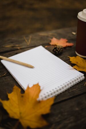 White blank notebook and pen on a dark background with a scarf, plaid and a cup, autumn yellow leaves and pine cones around. View from above. flat lay, in blur. Copy space.の写真素材