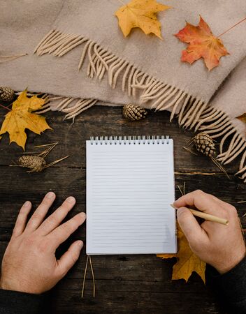 Human hands with a cup of coffee and scarf at wooden table with notebook and pen and autumn leaves. Human hands with a cup of coffee and scarf at wooden table.の写真素材