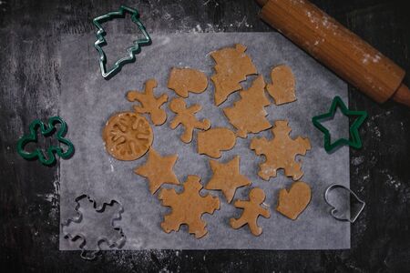 cut out gingerbread cookie in the form of a Christmas tree, star, little man, hearts from raw dough on parchment baking paper on a dark background. Top view. save space.の写真素材