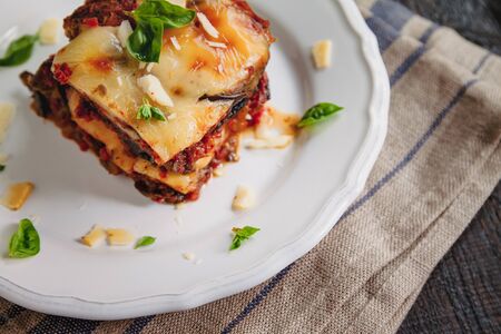 tradicional Parmigiana di melanzane: baked eggplant - italy, sicily cousine.Baked eggplant with cheese, tomatoes and spices on a white plate. A dish of eggplant is on a wooden tableの写真素材