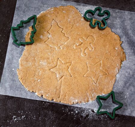cut out gingerbread cookie in the form of a Christmas tree, star, little man, hearts from raw dough on parchment baking paper on a dark background. Top view. save space.の写真素材