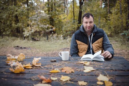 a young man sits in an autumn park at an old wooden table, drinks tea and reads a book. Warm dark autumn atmosphere, soft focus. In blur, copy space.の写真素材