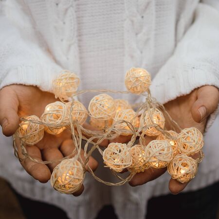 lights in the palms. Female hands hold a garland with wicker round balls. Girl in a white sweater with Christmas lights in her hands, warm New Year mood, soft focus.の写真素材