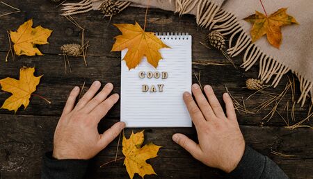Hands and White blank notebook with wooden letters phrase Good day on a dark background with a scarf, plaid and cup, autumn yellow leaves and pine cones around. View from above. flat lay, in blur. Copy space.の写真素材