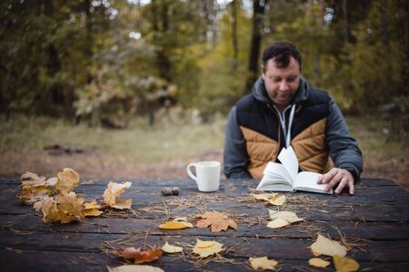 a young man sits in an autumn park at an old wooden table, drinks tea and reads a book. Warm dark autumn atmosphere, soft focus. In blur, copy space.の写真素材
